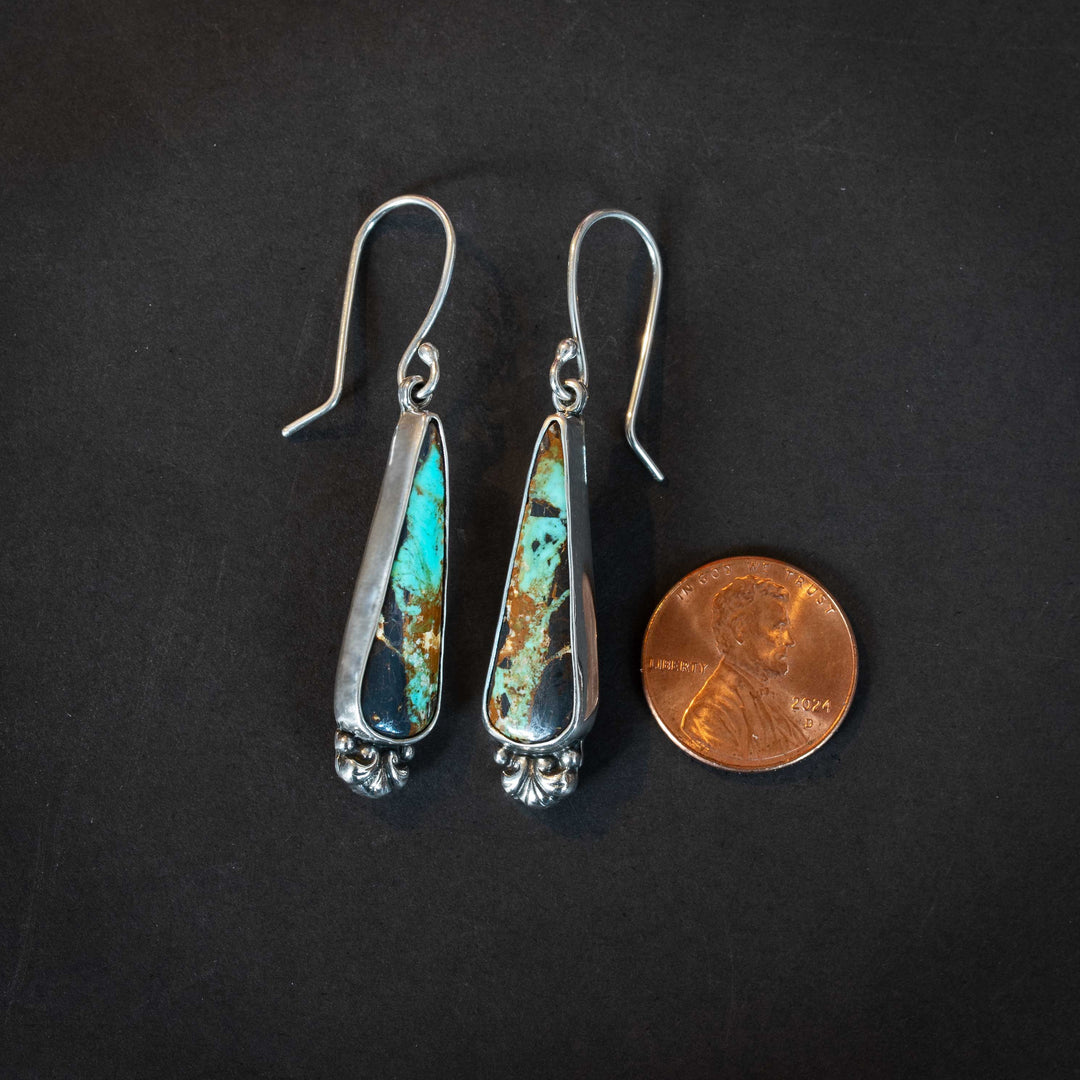 Black Jack turquoise sterling silver earrings shown next to a penny for scale, featuring elongated teardrop stones with rich brown matrix and turquoise veining.