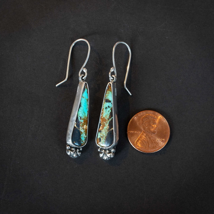 Black Jack turquoise sterling silver earrings shown next to a penny for scale, featuring elongated teardrop stones with rich brown matrix and turquoise veining.