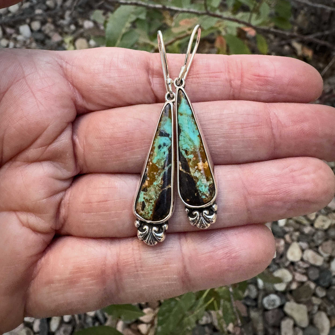 Pair of handmade Black Jack turquoise sterling silver earrings held in hand, showing natural stone color variation and delicate Art Nouveau silver detailing.