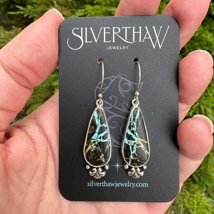 Handmade Blackjack turquoise earrings on earring card held outdoors, sterling silver French hooks, bold teal and gold veining in near-black chert matrix
