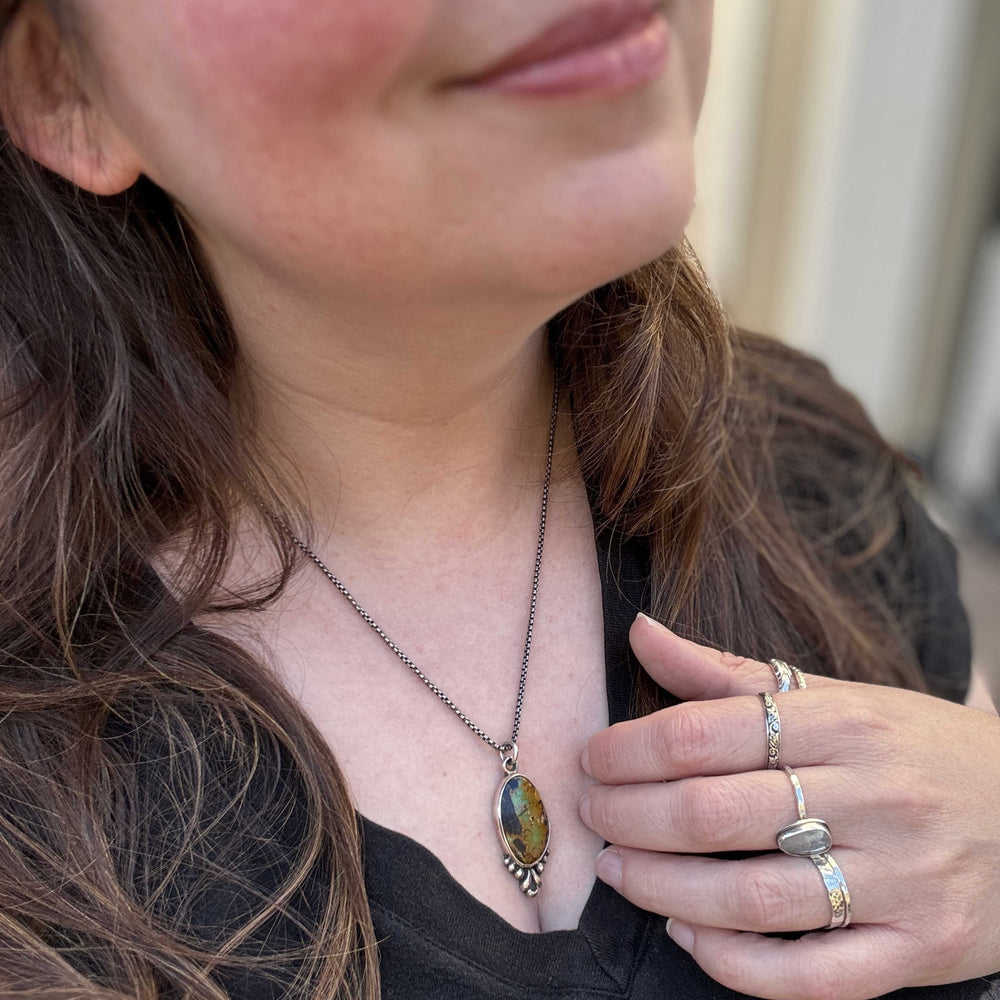 Close-up of a person wearing a necklace with an oval blackjack turquoise pendant, holding their hand near their face.