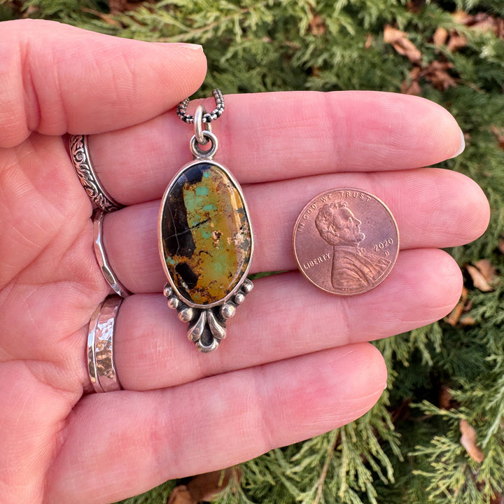 Hand holding a small silver pendant with a blackjack turquoise stone against a green leafy background