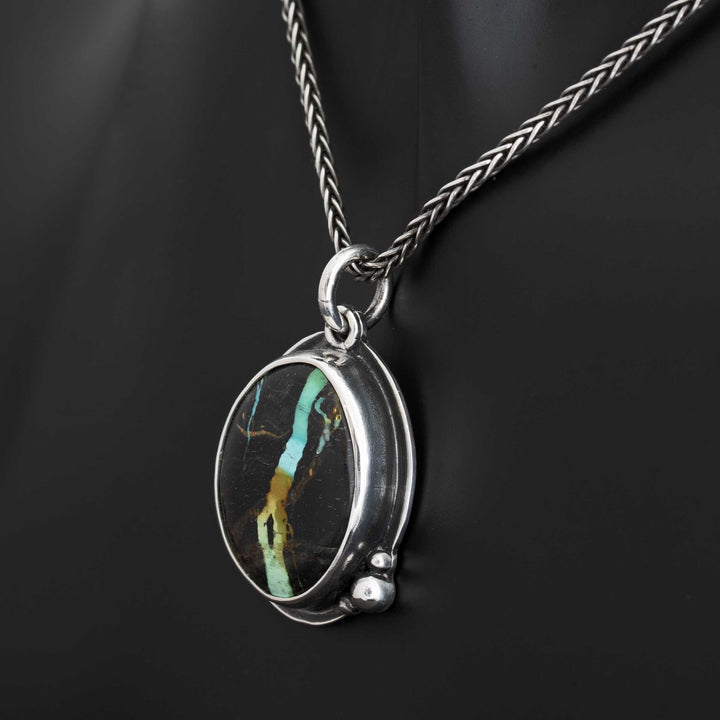 Angled view of Blackjack turquoise pendant on oxidized wheat chain against dark background, showing oval bezel setting and granule details