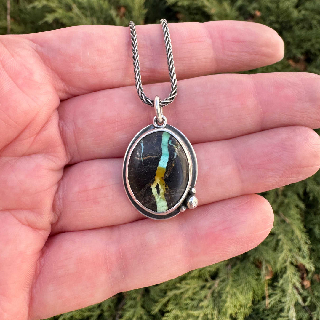 Handmade Blackjack turquoise pendant necklace in sterling silver, held outdoors against evergreen background — vivid teal and gold ribbon matrix in dark chert