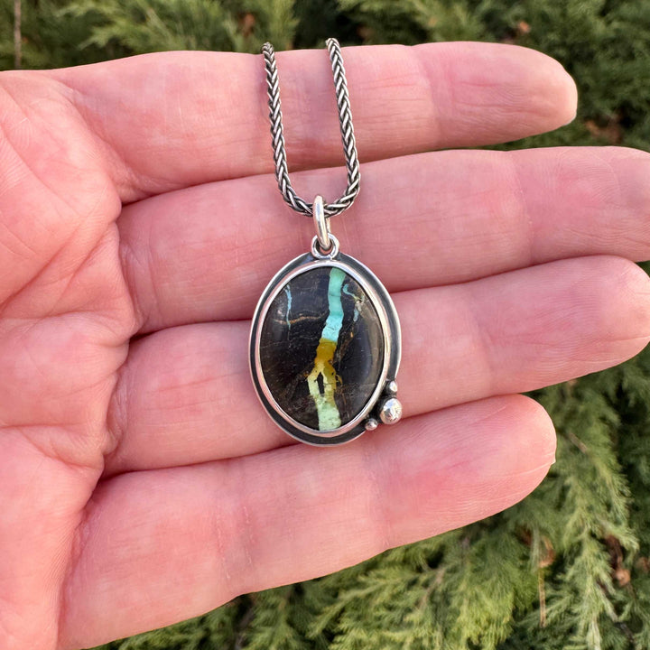 Handmade Blackjack turquoise pendant necklace in sterling silver, held outdoors against evergreen background — vivid teal and gold ribbon matrix in dark chert