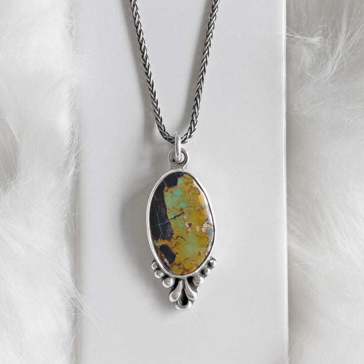 A sterling silver pendant necklace with a Blackjack turquoise stone, featuring a floral motif and a textured leaf-like back, displayed on a white furry surface.