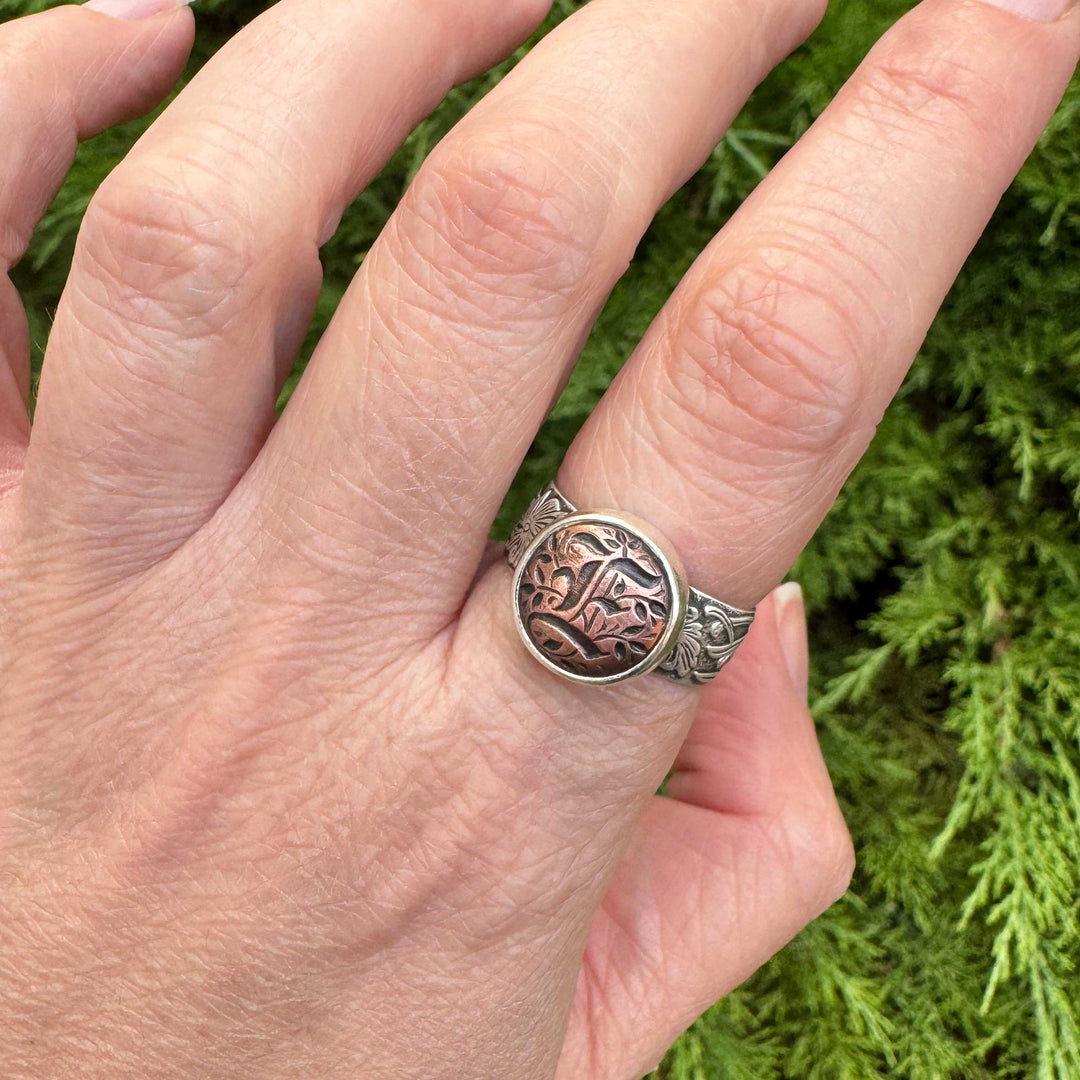 Gothic initial ring with a copper blackletter E worn on a hand, featuring a sterling silver floral band.
