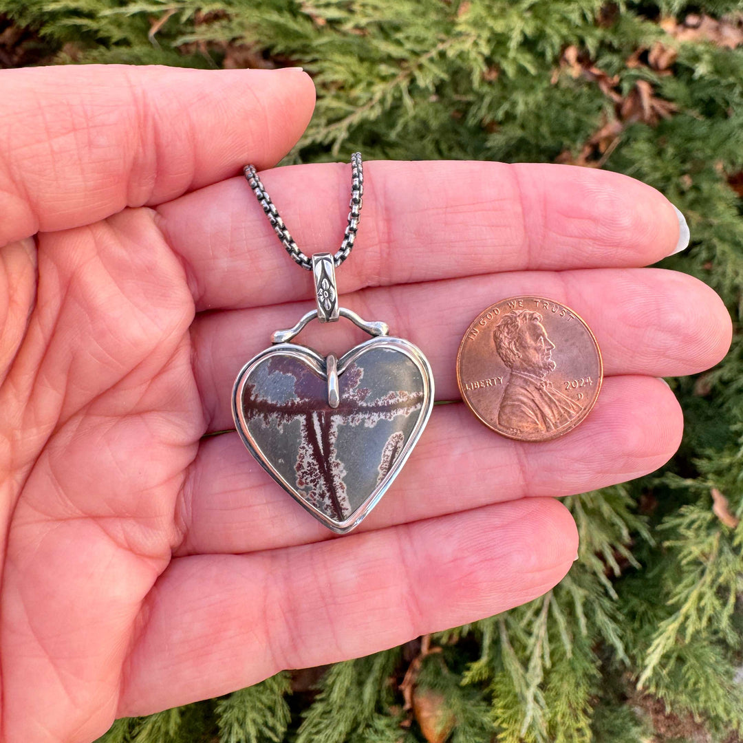 Heart-shaped pendant held in a hand with a penny for scale, against a green outdoor background.