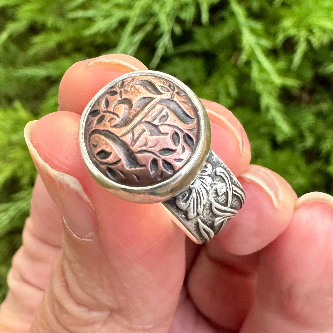 Handmade Gothic initial ring featuring a copper blackletter E set in a sterling silver stepped bezel with a floral patterned band.