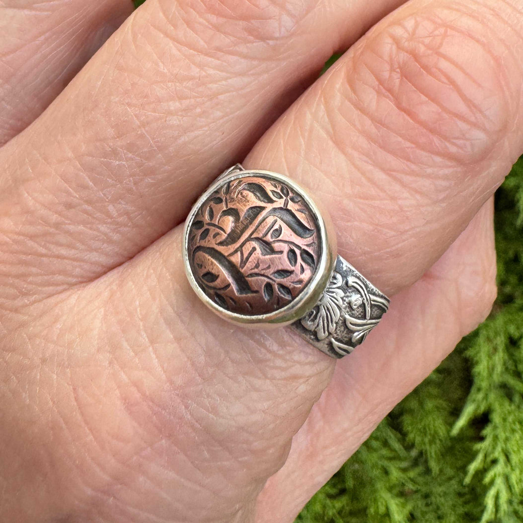 Angled close-up of a handmade sterling silver Gothic initial ring with a copper blackletter E centerpiece.