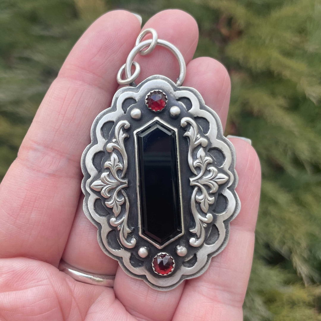 Handheld close-up of a gothic vampire statement necklace in sterling silver with black onyx and Mozambique garnets, showing ornate Victorian-inspired scrollwork