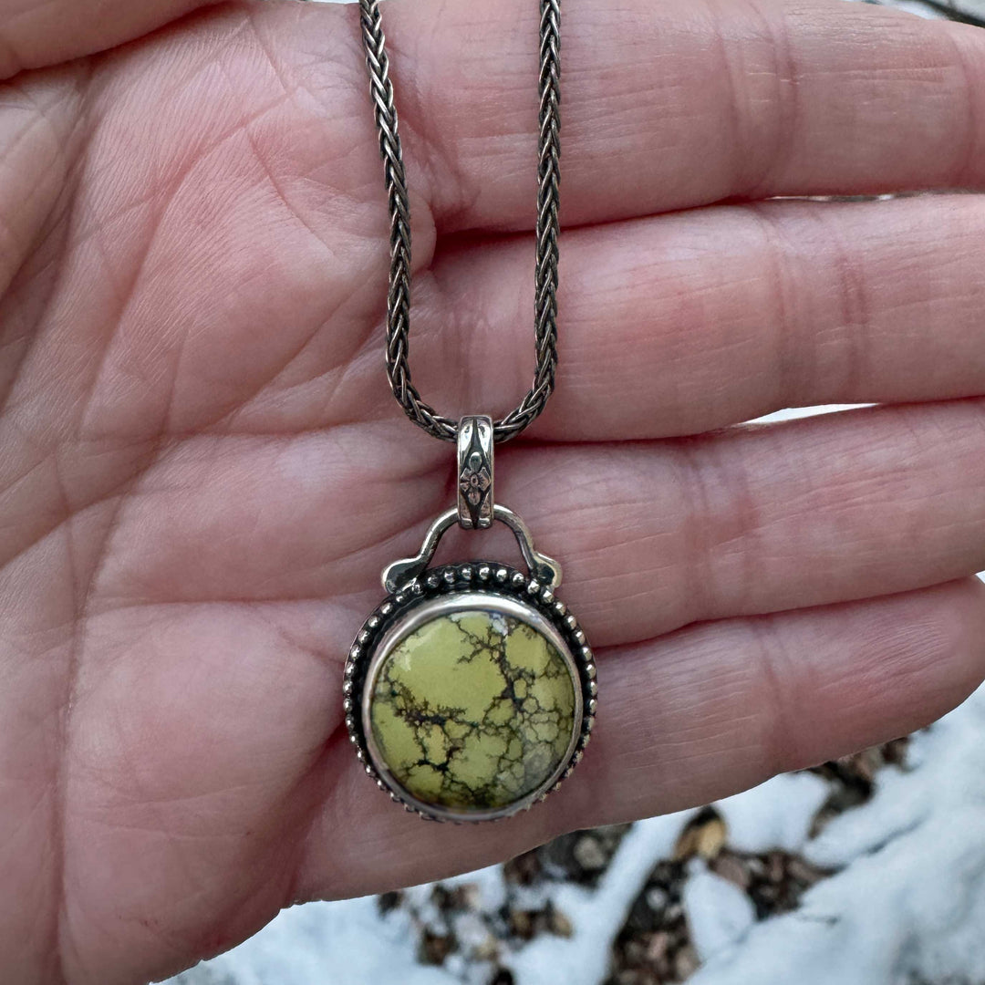 Round turquoise pendant on a silver chain photographed outdoors against snow and rocks.