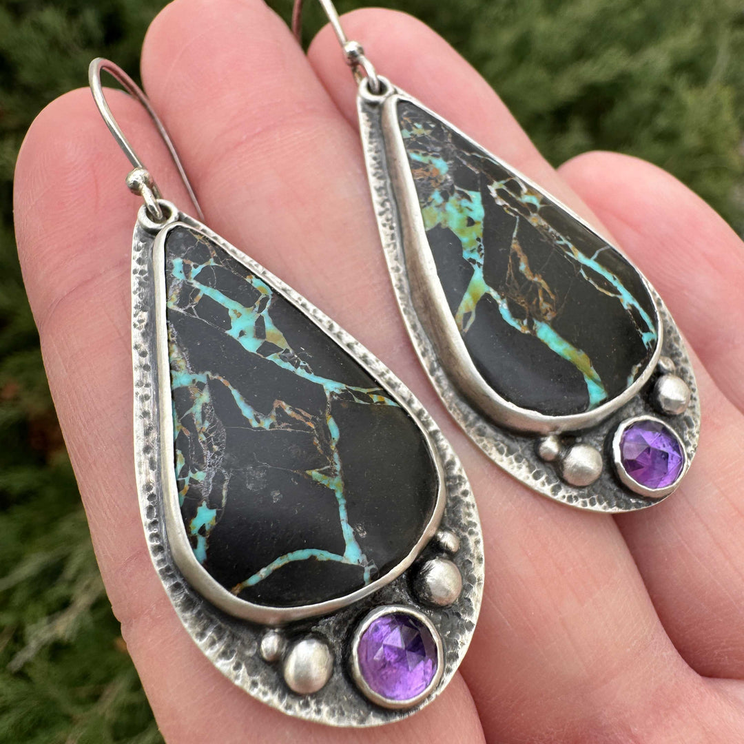 Close up of handmade Blackjack turquoise earrings with burr textured sterling silver frame and rose cut amethyst accents made in Colorado