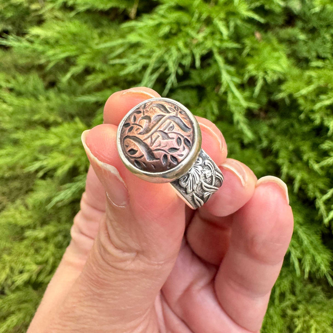 Handmade Gothic initial ring held between fingers, showing the copper blackletter E and sterling silver floral band.