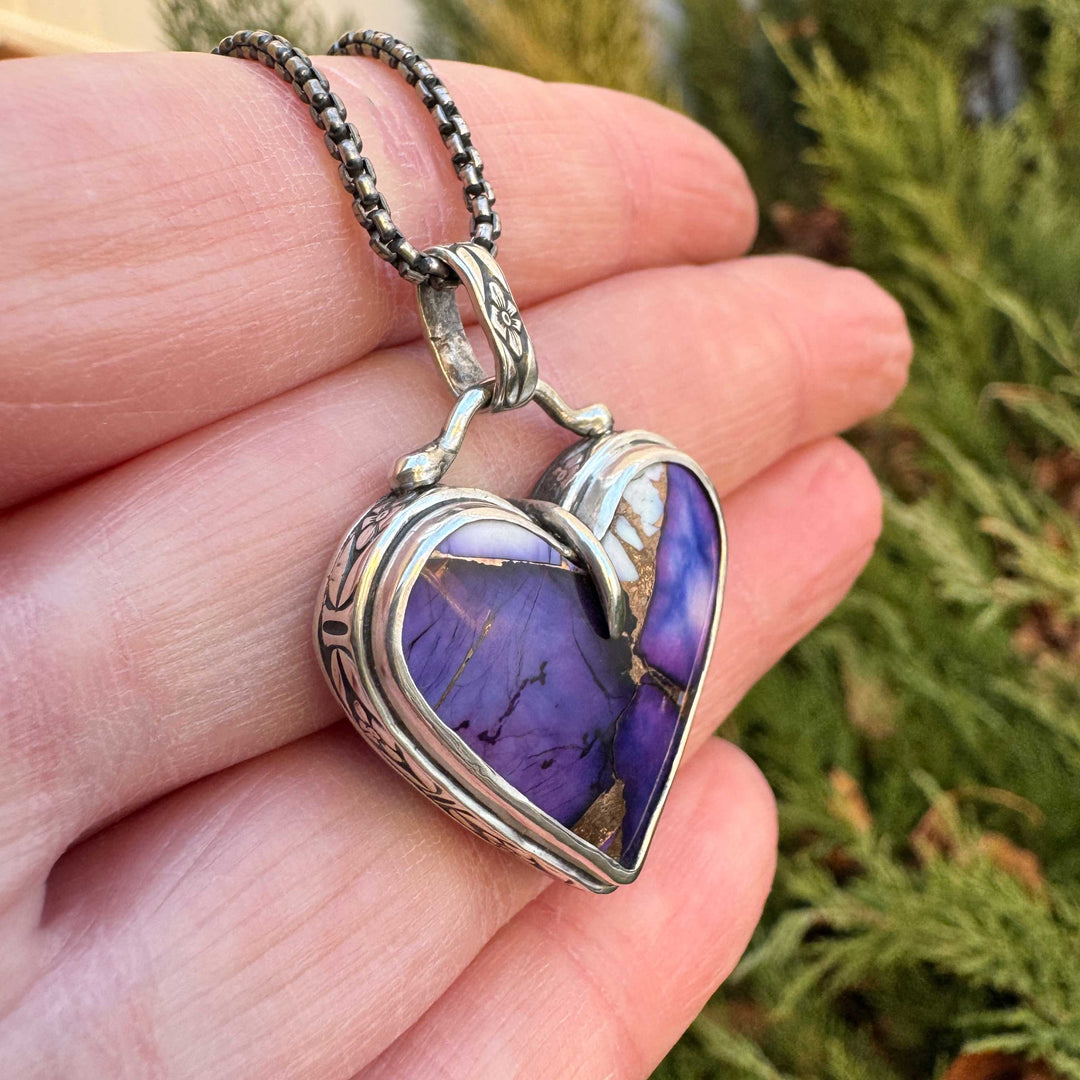 Heart-shaped purple stone pendant on a chain held in a hand with a blurred green background
