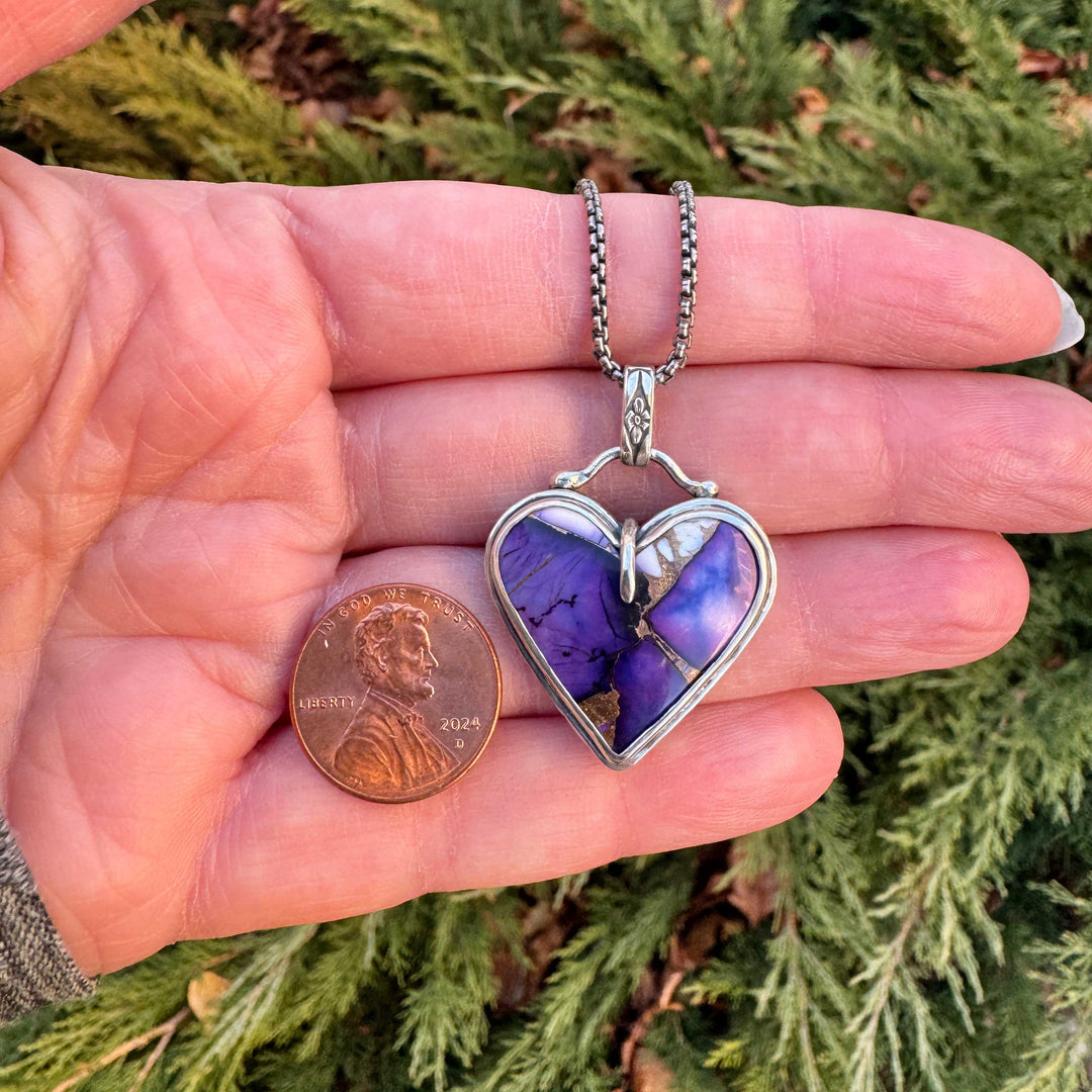 Heart-shaped purple pendant on a chain held between fingers with a penny for scale, against a green foliage background.