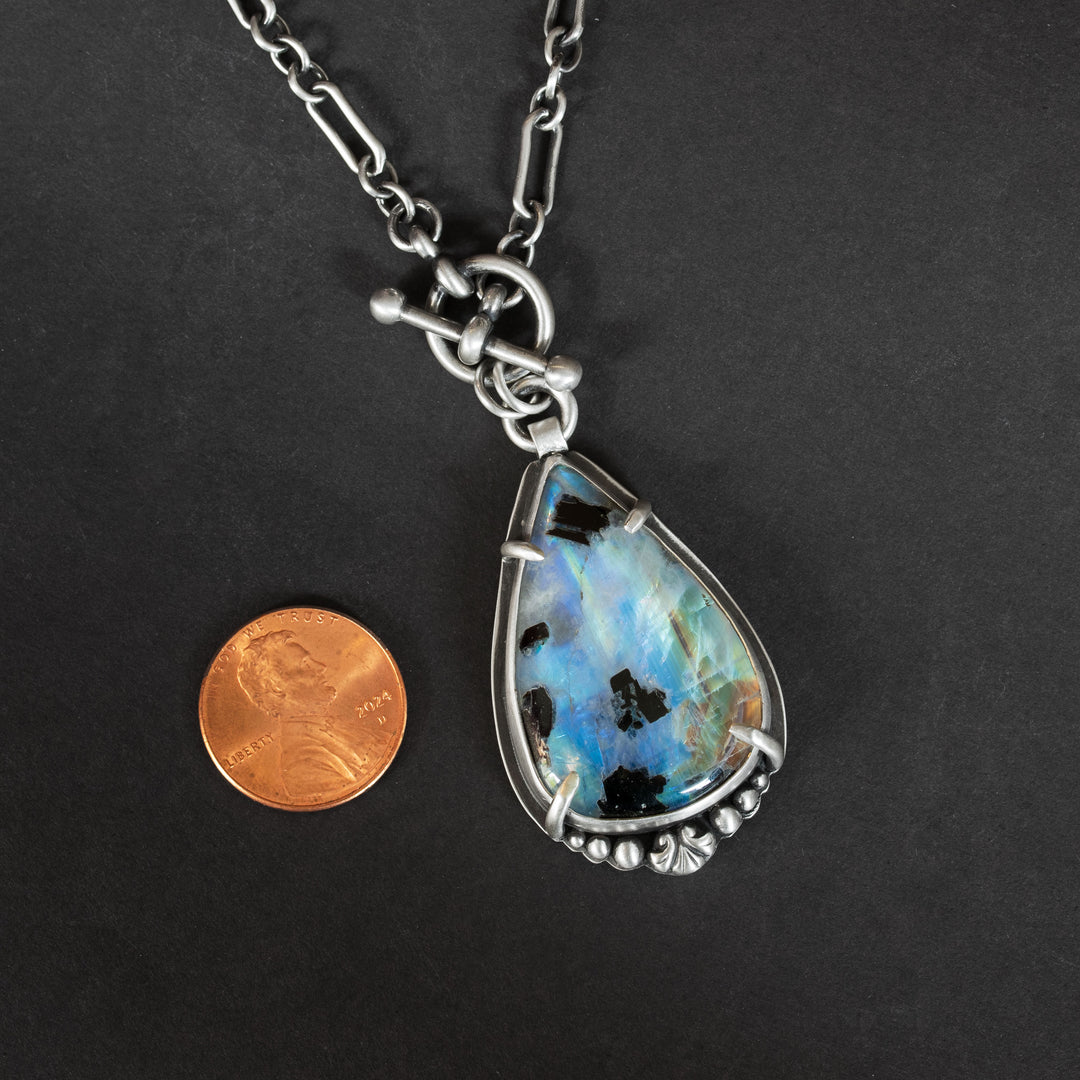 Rainbow moonstone and black tourmaline pendant on black background with penny for size reference.