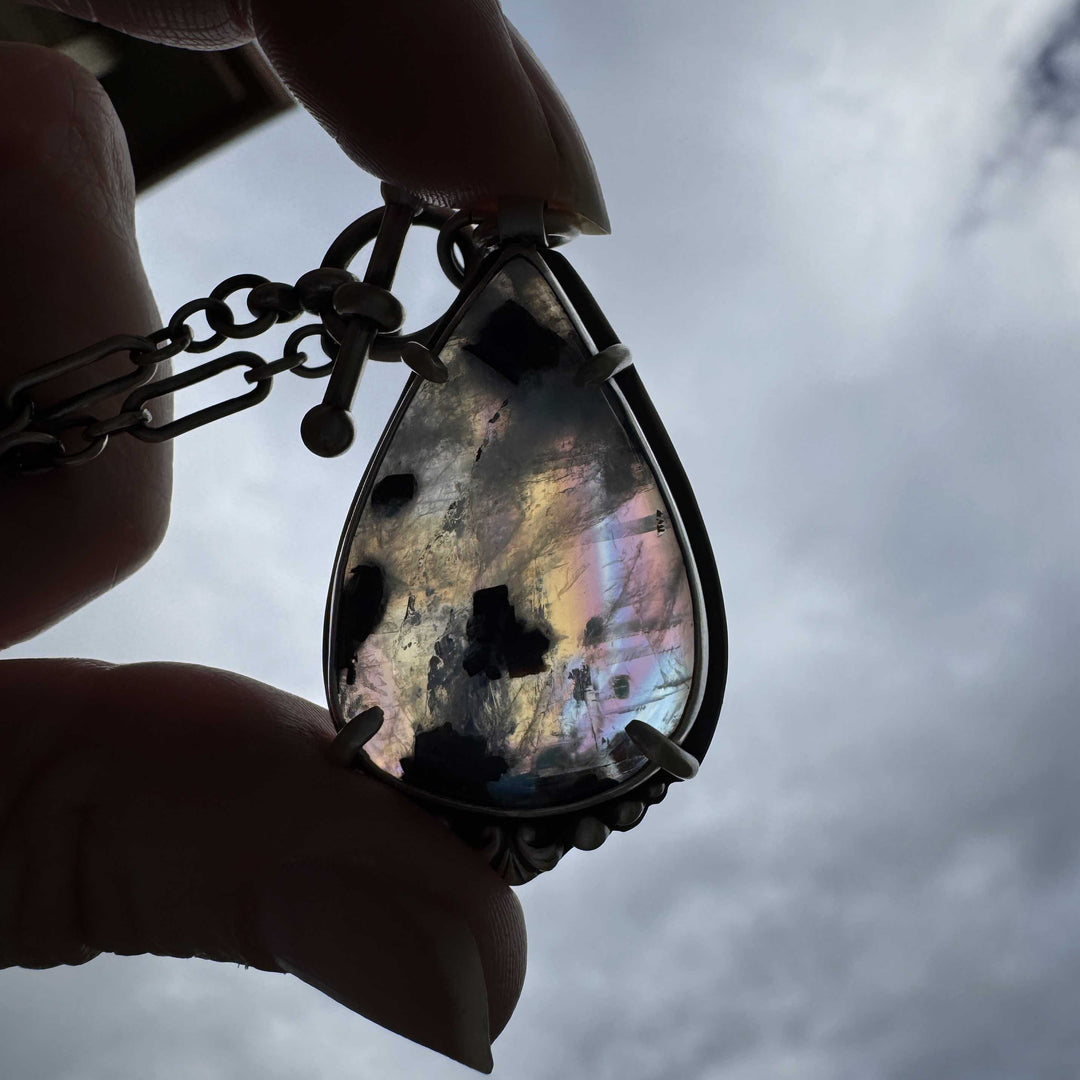 Teardrop-shaped rainbow moonstone pendant with rainbow-like iridescence held against a cloudy sky.