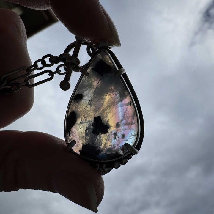 Teardrop-shaped rainbow moonstone pendant with rainbow-like iridescence held against a cloudy sky.