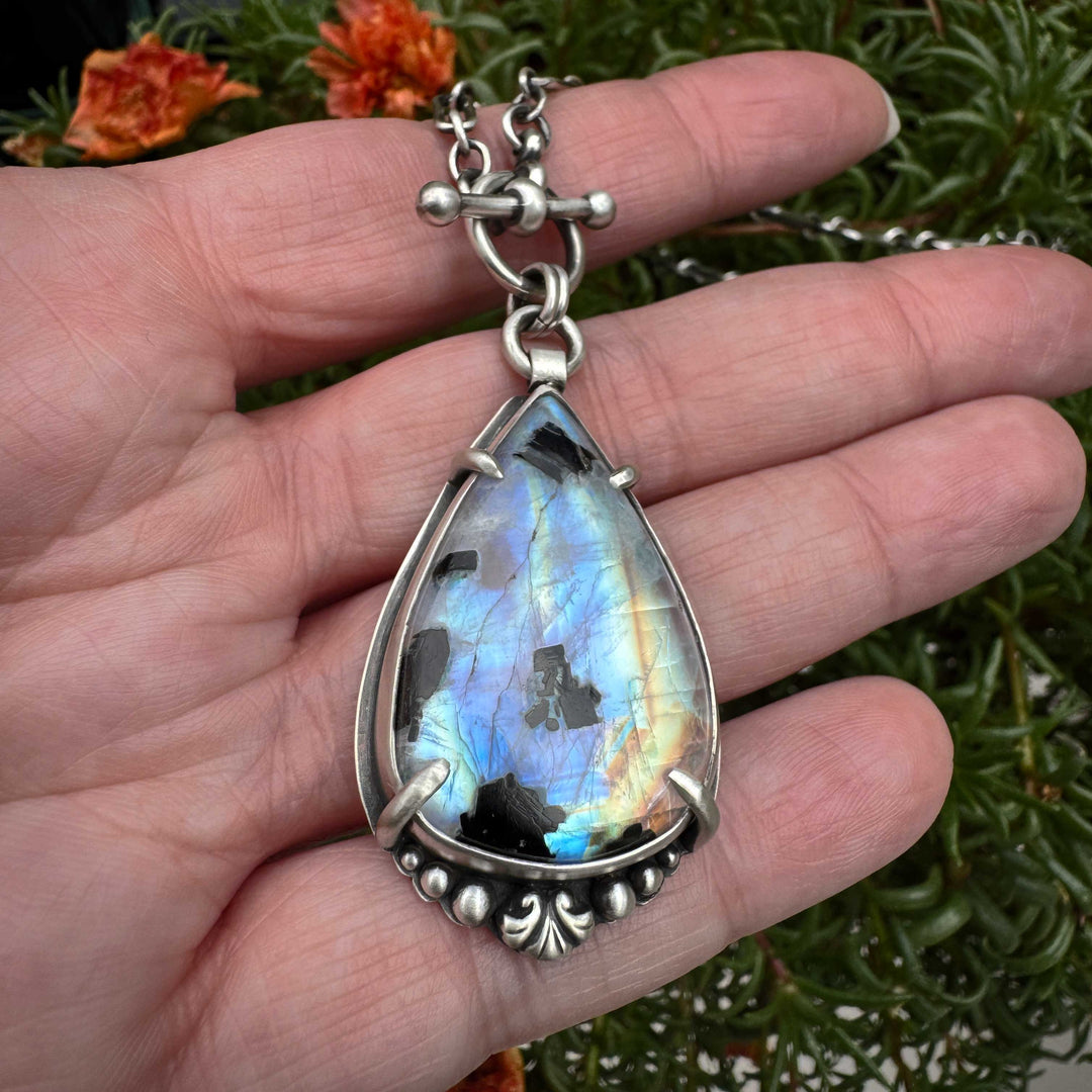 Teardrop-shaped rainbow moonstone pendant with silver frame held in a hand against a natural background
