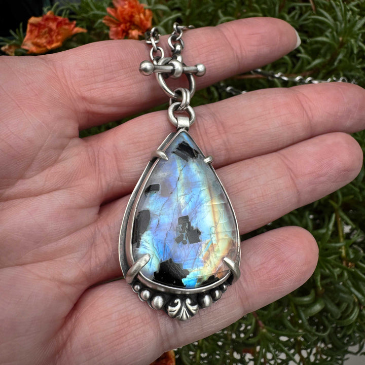 Teardrop-shaped rainbow moonstone pendant with silver frame held in a hand against a natural background