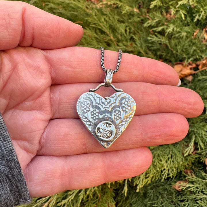 Textured back view of a heart-shaped silver pendant held in a hand with a green outdoor background