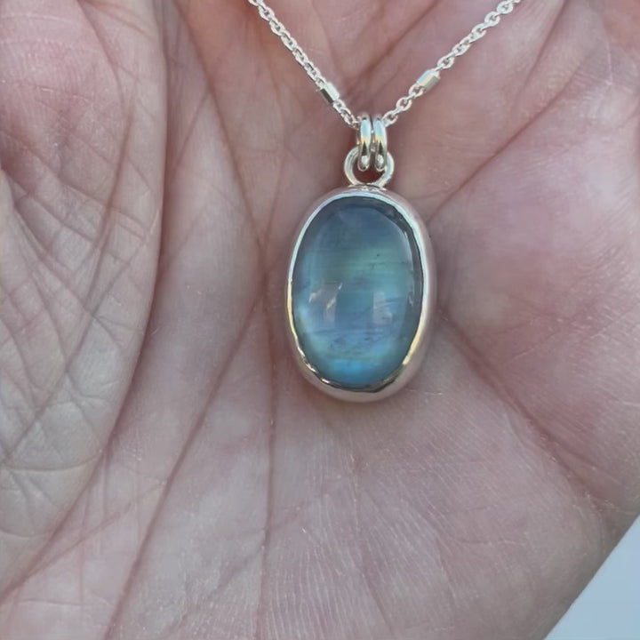 A video of a rainbow moonstone pendant necklace in sterling silver. The stone glows blue and green. 