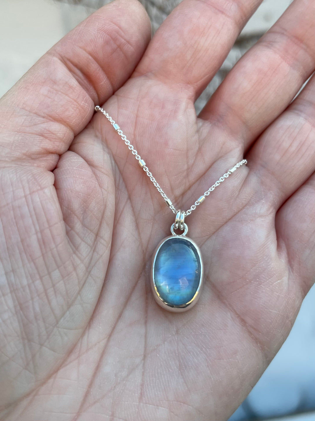 A hand holding a rainbow moonstone pendant necklace in sterling silver. Made in northern Colorado. 